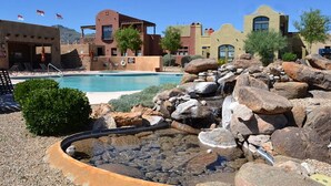 Una piscina con cascata artificiale rocciosa, circondata da edifici in stile desertico con tetti piani e ampie finestre.