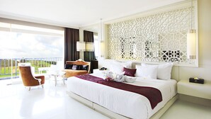 A modern hotel room with a large bed, a balcony with a view, a sitting area with a chair and table, and a decorative wall panel. - The Rich Prada Bali (Pecatu)