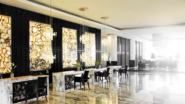 A modern hotel lobby with marble flooring, a reception desk, black chairs, and a large window with a decorative wall. - The Rich Prada Bali (Pecatu)