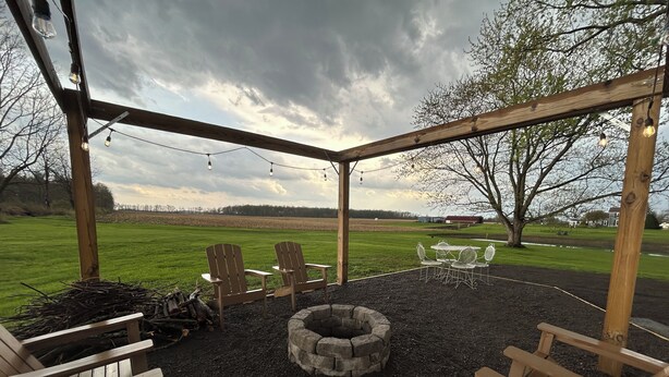 Ein Sitzbereich im Freien mit Adirondack-Stühlen und einer Feuerstelle aus Stein, umgeben von einer hölzernen Pergola und Lichterketten.