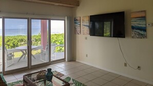Habitación con un amplio ventanal corredero que da a un balcón con vistas al océano. Hay un televisor de pantalla plana montado en la pared, y una mesita con un jarrón y una cesta.