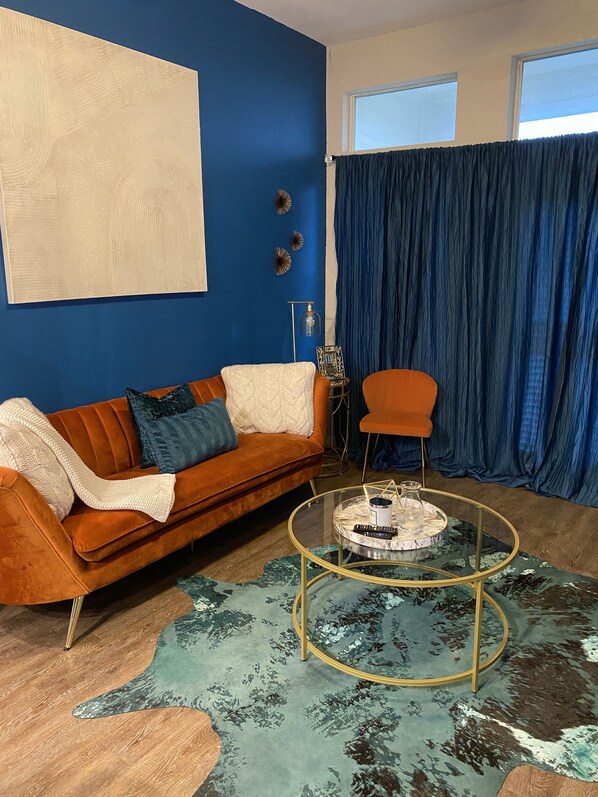 A living room with a blue accent wall, a brown sofa with patterned cushions, a glass coffee table, and a blue curtain.