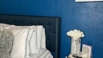 A bedroom with a dark blue wall, a bed with white bedding, a small bedside table with a vase of white flowers, and decorative wall art.