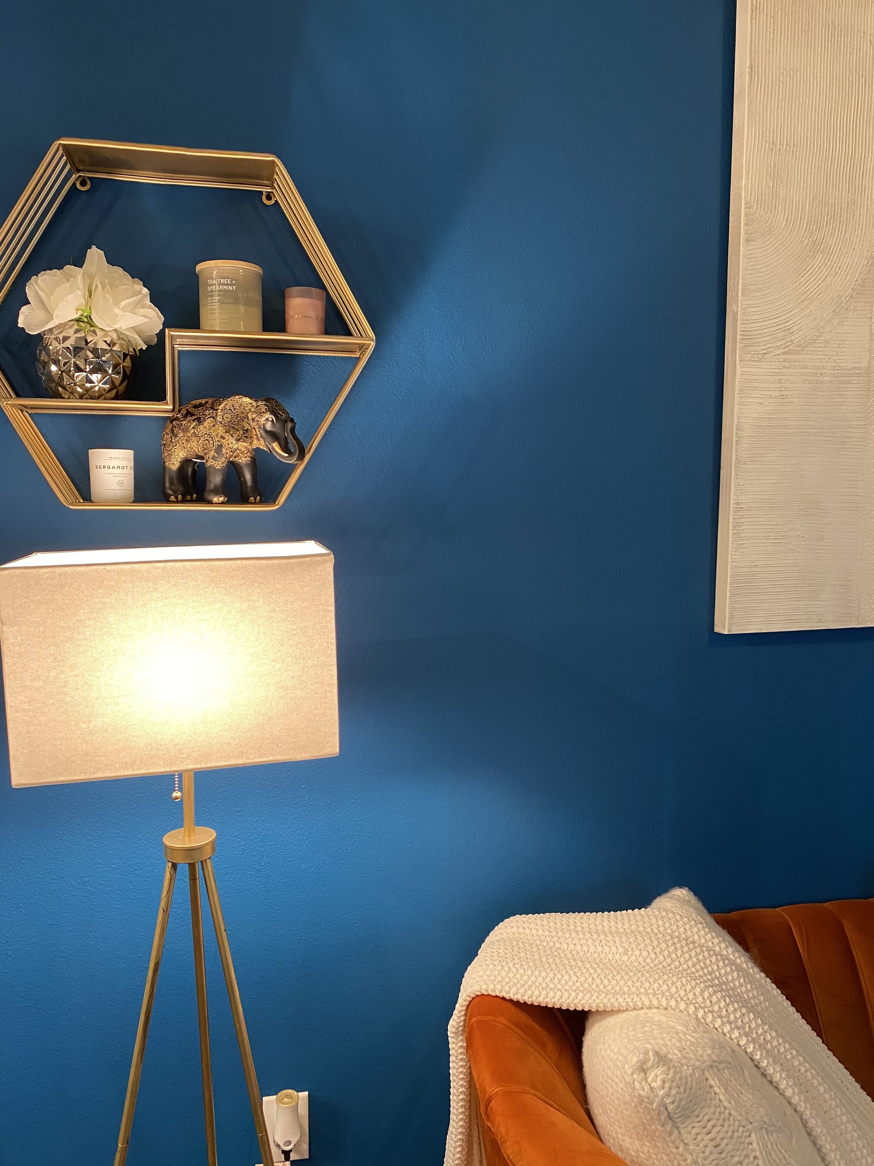 Une pièce avec un mur bleu, un miroir hexagonal, une lampe et un lit recouvert d’une couverture blanche.