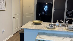 A modern kitchen with a marble countertop, a black refrigerator, a coffee bar sign, and a teal chair. - Sophisticated Haus- Houston Near NRG Stadium (Houston)