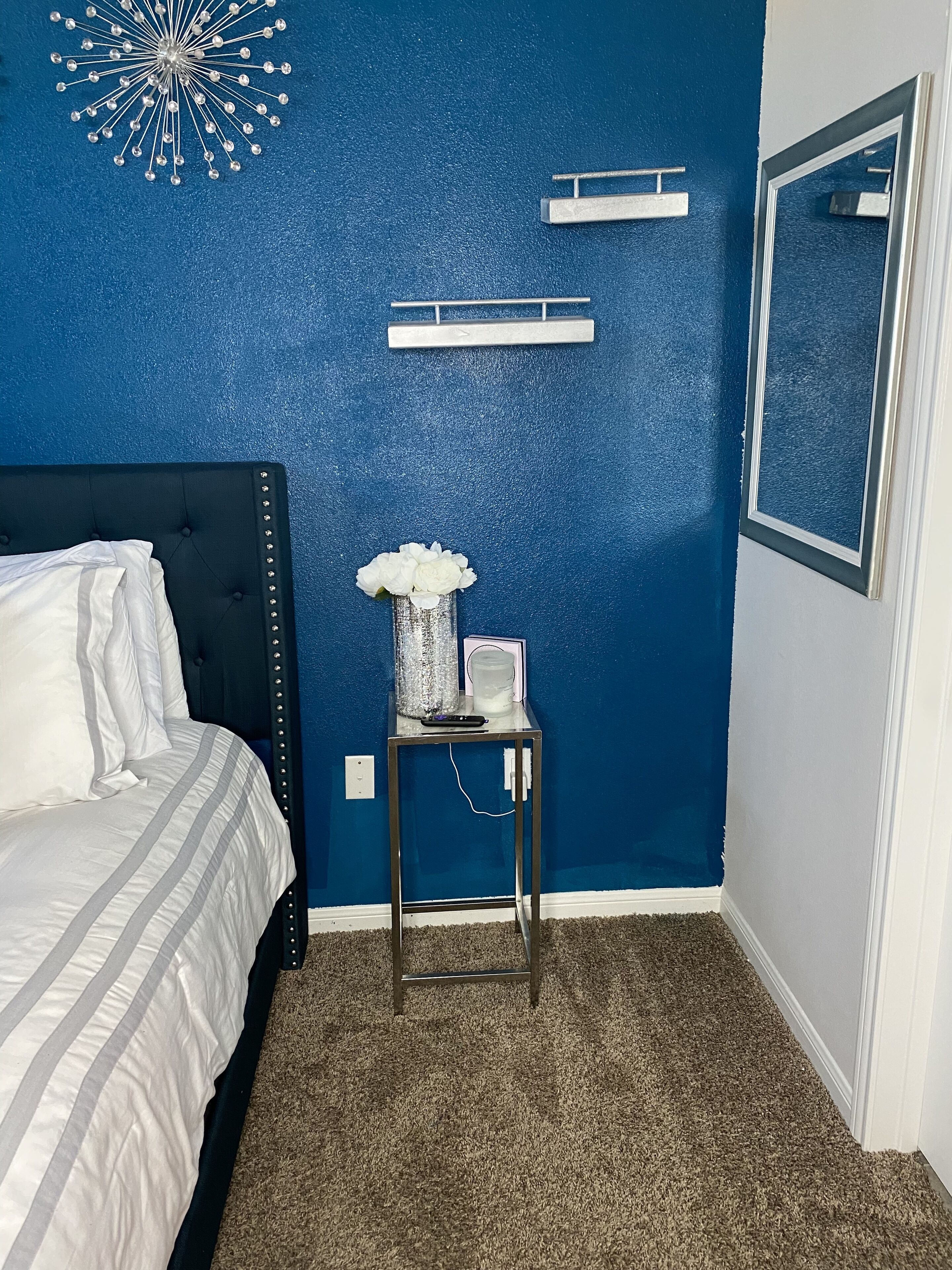 A bedroom with a bed, a small table with a vase, and a blue wall with a decorative item.