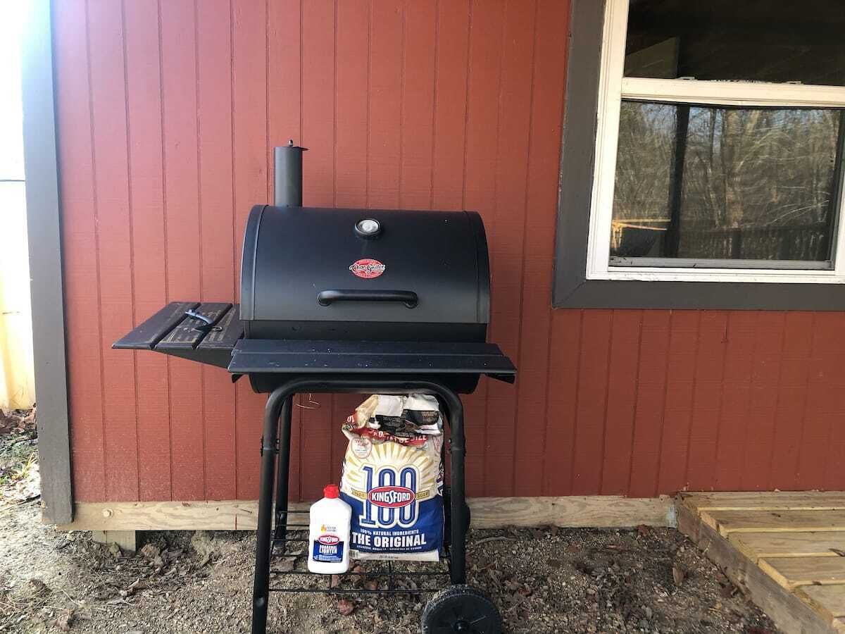 Un barbecue avec un sac de charbon de bois Kingsford et une bouteille d’huile de cuisson, devant un bâtiment rouge.