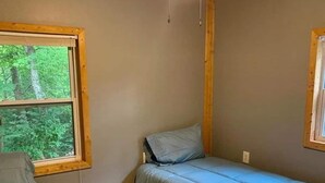 A small, single-room cabin with a wooden floor, two beds, a window with a view of trees, and a simple wooden ceiling.