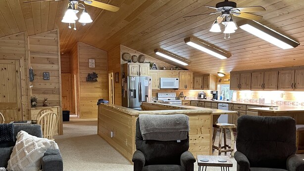 A cozy cabin interior with a living area featuring a sofa and armchairs, a kitchen with wooden cabinets and appliances, and a ceiling fan. - Adventure Basecamp near YNP • Hot Tub + Toy Garage (Island Park)