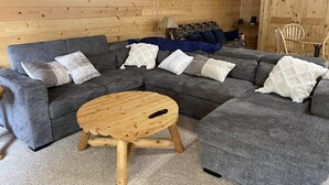 A cozy log cabin interior with a grey sectional sofa, a round wooden coffee table, and a dining area with a wooden table and chairs. - Adventure Basecamp near YNP • Hot Tub + Toy Garage (Island Park)