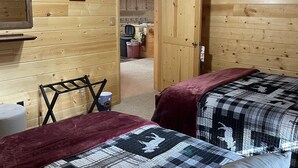 A cozy cabin room with two beds, a ceiling fan, wooden walls, a kitchen area in the background, and a framed picture on the wall. - Adventure Basecamp near YNP • Hot Tub + Toy Garage (Island Park)