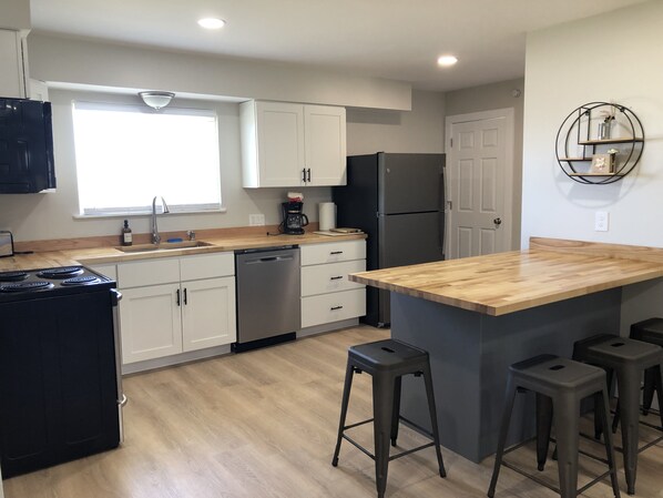 A modern kitchen with white cabinets, a black refrigerator, a coffee maker, and a wooden island with stools. - Fully Renovated Peek-A-Boo Bayview Duplex-10 minuets from Downtown Traverse City (Williamsburg)