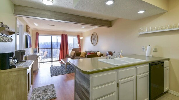 A modern kitchen with a large island, white cabinetry, and a dishwasher. There is a dining area with a table and chairs, and a view of the outdoors through a sliding glass door. - Mountain Getaway- Outdoor Pool! Hiking Trails! (Reeds Spring)