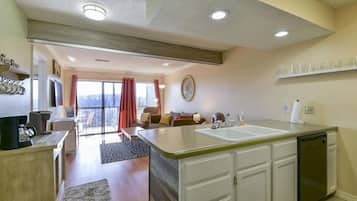 A modern kitchen with a large island, white cabinetry, and a dishwasher. There is a dining area with a table and chairs, and a view of the outdoors through a sliding glass door.