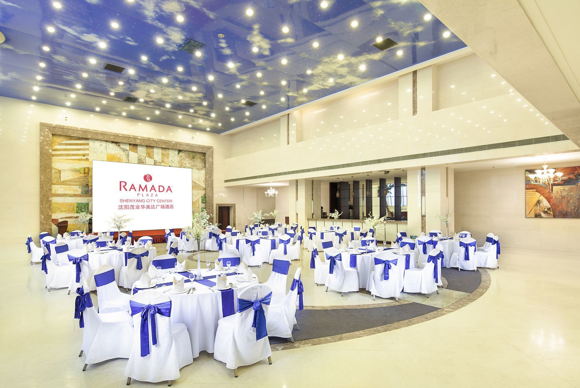 A spacious hall with rows of round tables, each with white tablecloths and blue sashes, set up for an event.