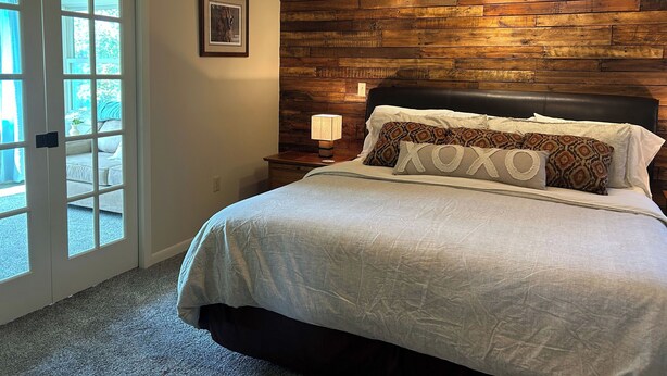 A bedroom with a wooden headboard, a bed with white bedding, a nightstand with a lamp, and a door leading to an outdoor area. - Serenity Heights - Peaceful Country Acres with beautiful view in every direction (Osceola)