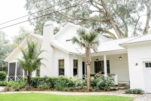 A white house with a porch, surrounded by greenery and trees.