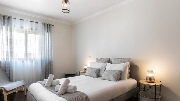 A neatly arranged bedroom with a large bed, bedside tables with lamps, a chair, and a window with sheer curtains. - O Sítio do Areal - Bright House in Nazaré (Nazaré)