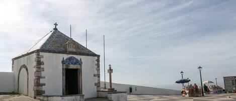 A small chapel with a tiled roof and a cross on top, located on a paved area with a tiled floor.