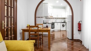 Ein Essbereich mit Holztisch und Stühlen, eine durch einen Bogen sichtbare Küche sowie ein Feuerlöscher an der Wand.