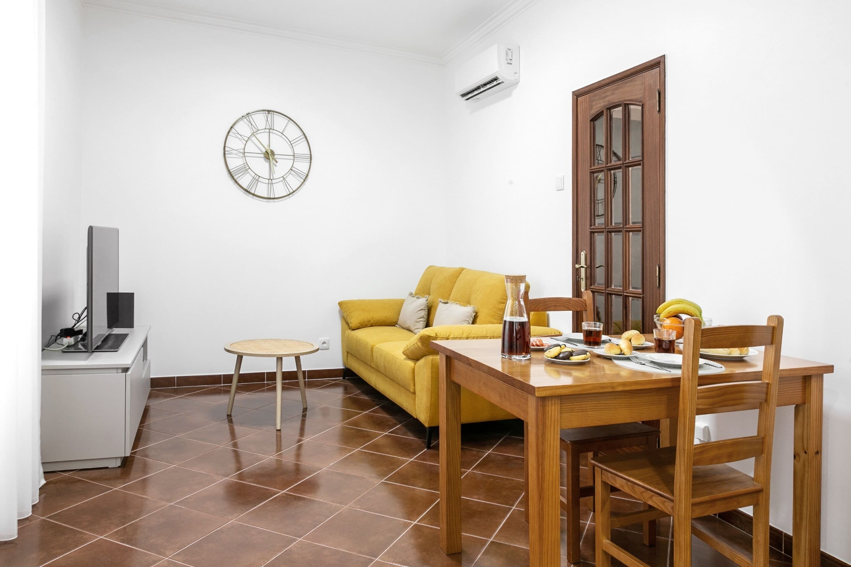 A compact living space with a dining table, a sofa, a small side table, a TV, and a clock on the wall.