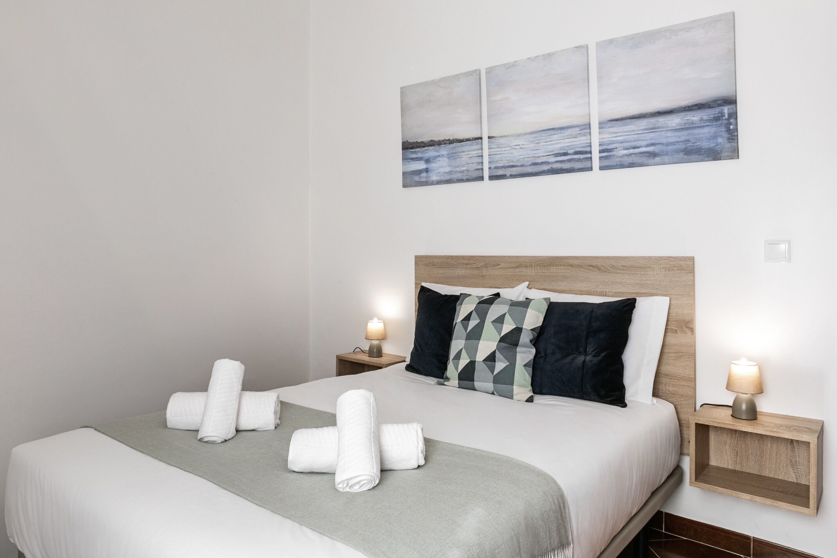 A neatly made bed with rolled towels, a grey throw blanket, and a headboard with geometric pillows. There are two bedside tables with lamps, and a large abstract painting above the bed.