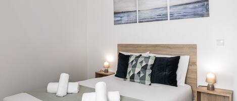 A neatly made bed with rolled towels, a grey throw blanket, and a headboard with geometric pillows. There are two bedside tables with lamps, and a large abstract painting above the bed.