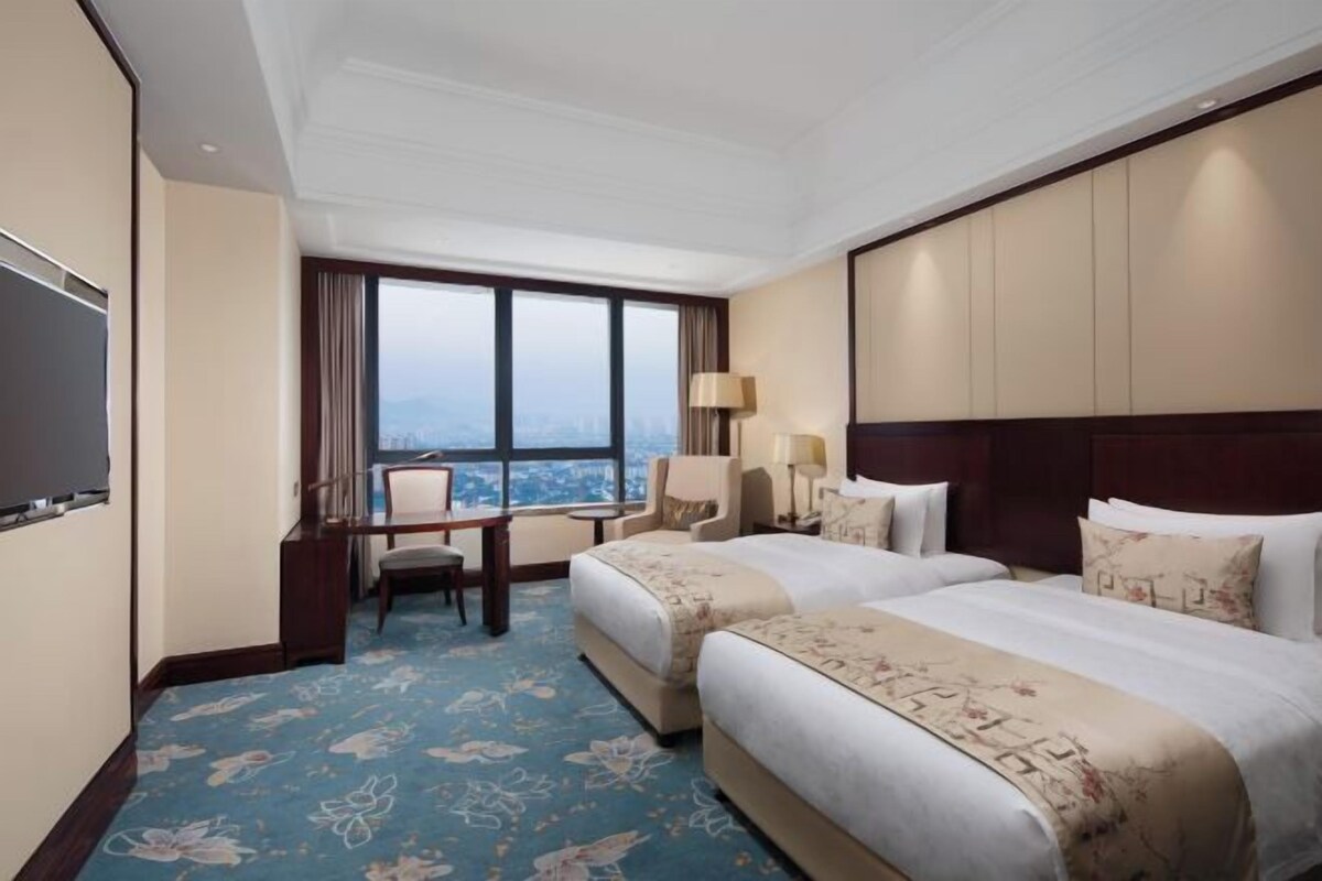 Habitación de hotel con dos camas, un escritorio, una silla y un amplio ventanal con vista a la ciudad.