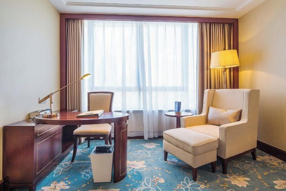 Habitación de hotel con escritorio, silla, sillón y un amplio ventanal con cortinas.