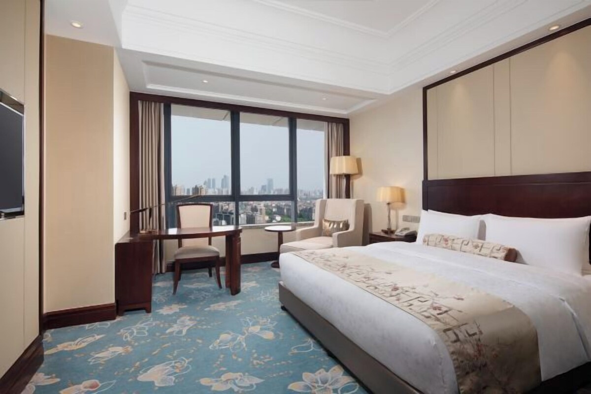 Habitación de hotel con una cama grande, un escritorio, una silla y vistas a la ciudad a través de amplios ventanales.