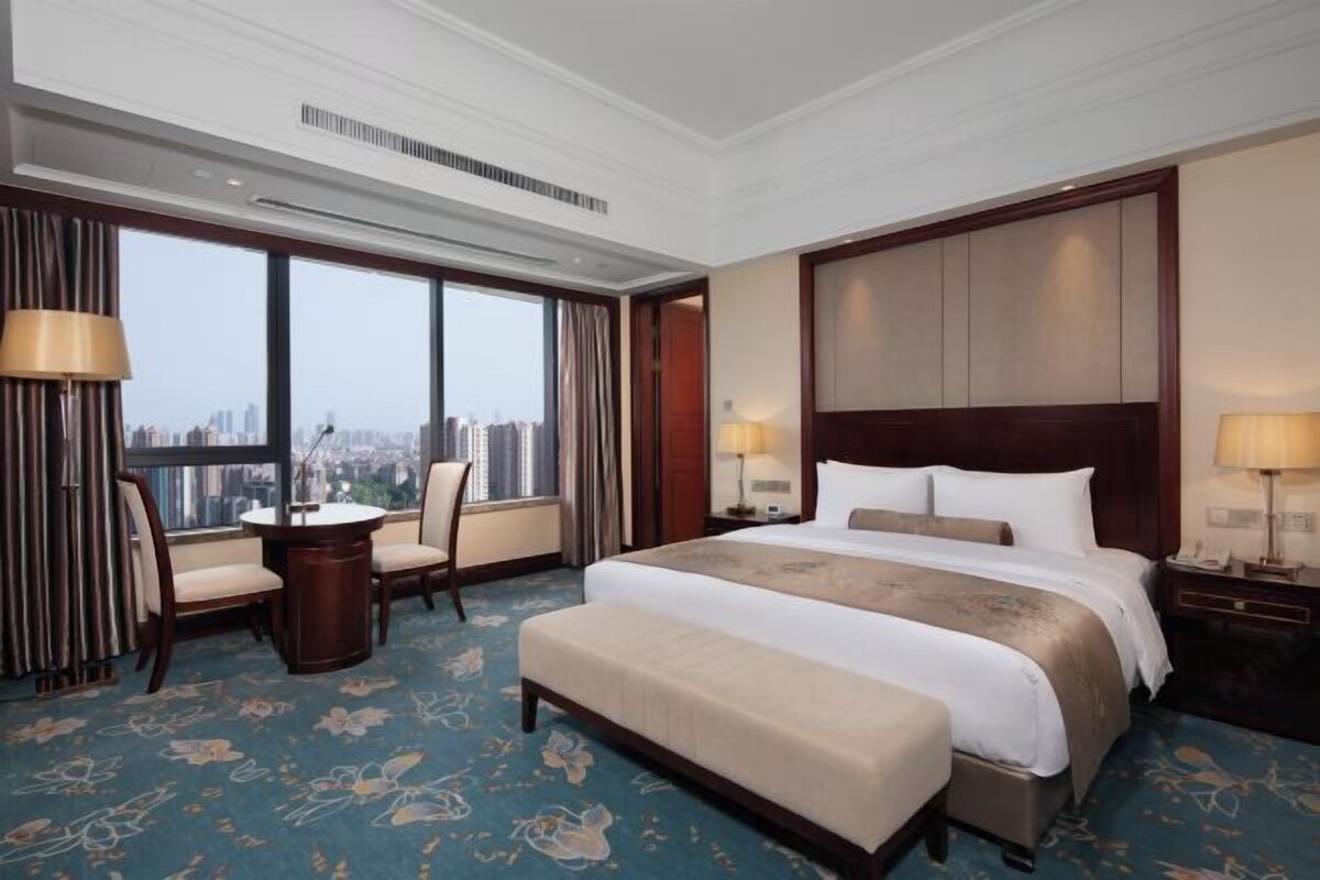 Habitación de hotel con una cama grande, un escritorio, dos sillas, vista a la ciudad y un ventanal amplio.