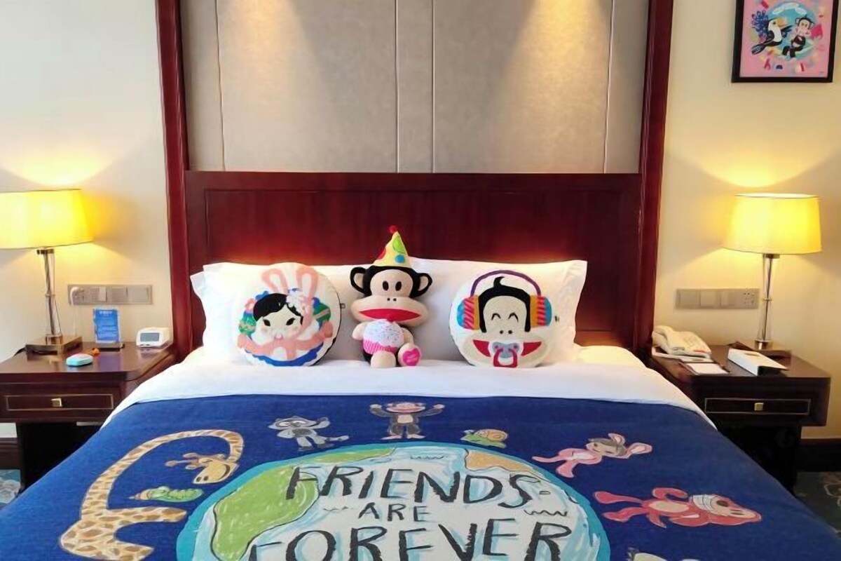 Habitación de hotel con una cama, un edredón y almohadas con temática de 'Friends' y personajes de caricatura. Hay dos mesitas de noche, lámparas y un reloj.