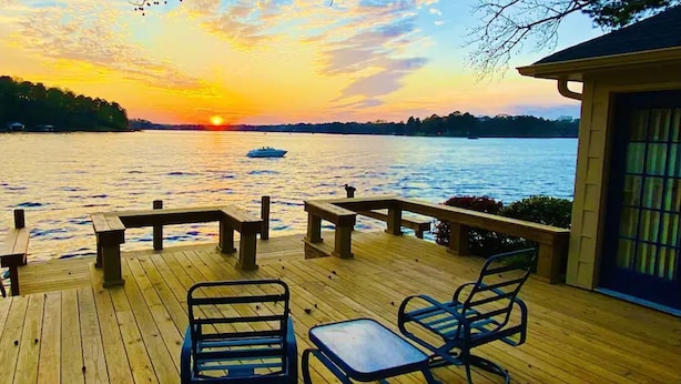 Terraza de madera con bancos y mesas, con vista a un cuerpo de agua al atardecer.