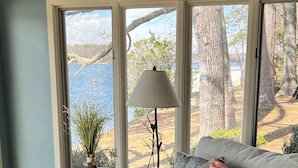 Una sala de estar con un sofá gris, una mesita redonda con una lámpara y vistas a una masa de agua a través de ventanas amplias.