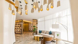 En modern hotellobby med ett marmorgolv, en soffa med kuddar, ett sidobord, en golvlampa och en bokhylla.