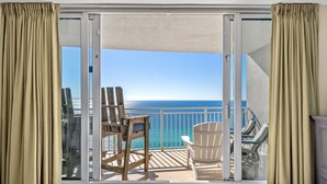 A balcony with a view of the ocean, a wooden chair, a white chair, and a glass door.
