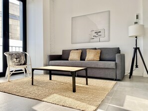 Un salon moderne avec un canapé gris, une table basse en bois, un fauteuil blanc orné d’un coussin à motif de lion, un tapis tissé et un lampadaire.