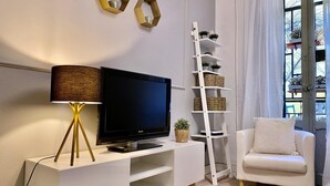 A modern living room with a flat-screen TV on a white stand, a floor lamp, a white sofa, a white shelving unit with baskets, and a window with curtains. - Malasaña Gallery – 3-Bedroom Apartment in the City Center (Madrid)