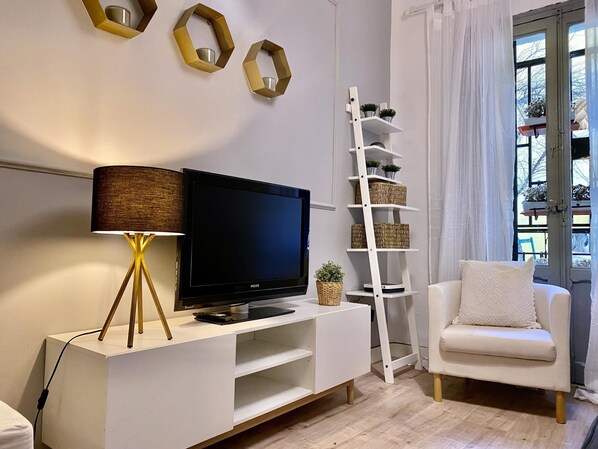 Un salon moderne avec un téléviseur à écran plat posé sur un meuble blanc, un lampadaire, un canapé blanc, une étagère blanche avec des paniers, et une fenêtre avec des rideaux.
