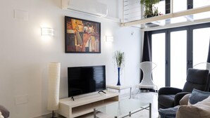 Ein modernes Wohnzimmer mit Flachbildfernseher, Glastisch und Sofa. An der Wand hängt ein Gemälde und auf einem Beistelltisch steht eine Topfpflanze.