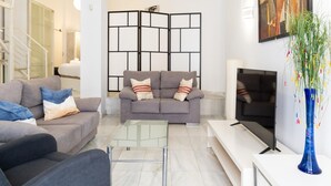 Ein modernes Wohnzimmer mit einem grauen Sofa, einem Glastisch, einem Flachbildfernseher und einer blauen Vase mit Blumen.