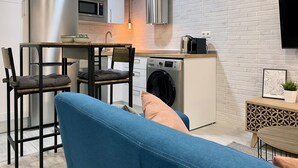 A compact kitchen with a stainless steel refrigerator, a microwave, and a washing machine. There is a dining area with a blue sofa and a small table. - Madrid Styles - One-bedroom apartment (Madrid)