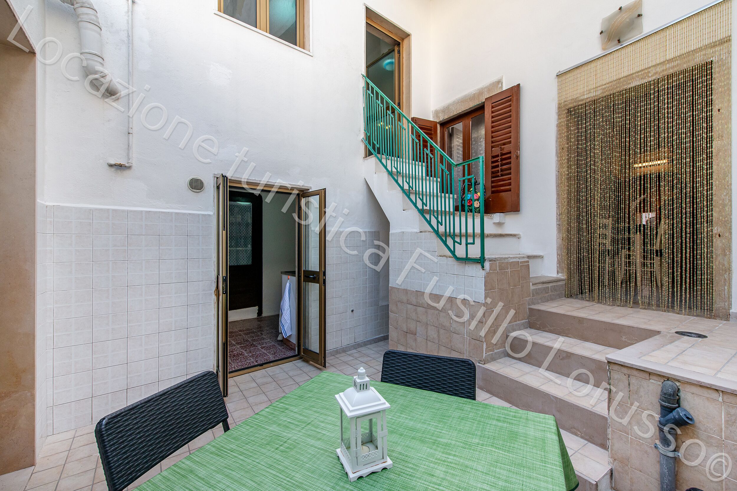 Un patio con una mesa verde, sillas, un farol, una escalera y una puerta que da a un espacio interior.