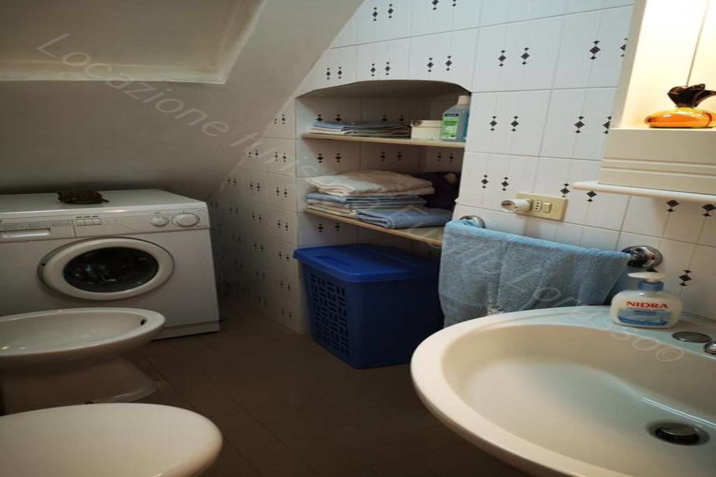 A compact bathroom with a washing machine, toilet, sink, and shelves with folded towels.