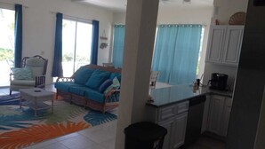 A room with a sofa, a small table, a kitchen area with cabinets and a coffee maker, and a view through a window with blue curtains.