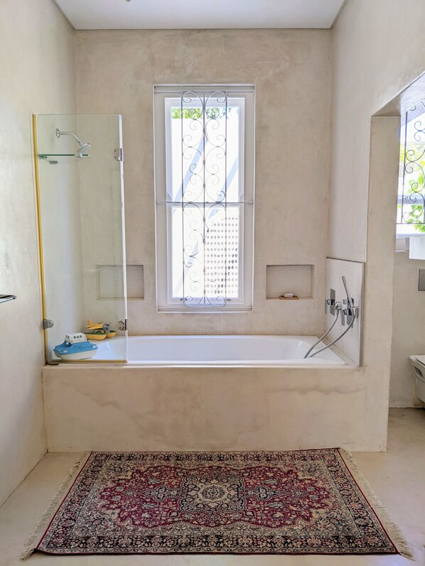 Une salle de bain avec une baignoire, une cabine de douche en verre, une fenêtre ornée de barreaux en métal décoratifs et un tapis à motifs sur le sol.