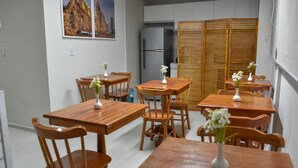 Uma área de refeições com mesas e cadeiras de madeira, vasos brancos com flores e uma geladeira ao fundo.