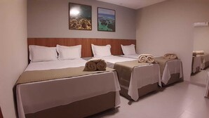Quarto de hotel com três camas, cada uma com roupa de cama branca e cabeceira marrom. Existem duas molduras com imagens na parede acima das camas.
