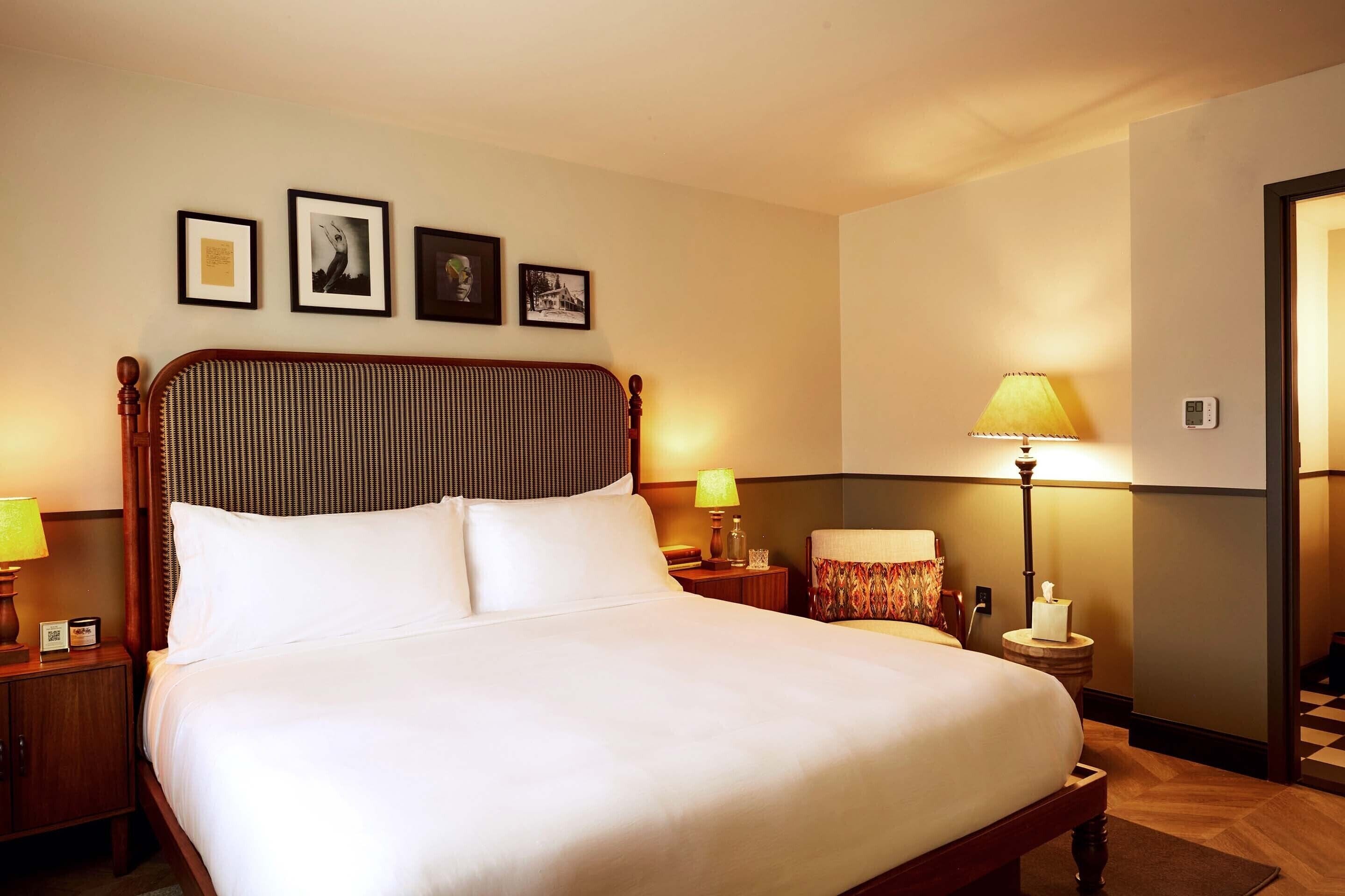 A neatly made bed with white linens, a wooden headboard, and a nightstand with a lamp. There are framed pictures on the wall above the bed.