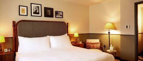 A neatly made bed with white linens, a wooden headboard, and a nightstand with a lamp. There are framed pictures on the wall above the bed.